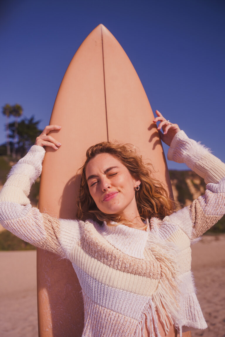 Woman with curly hair wearing a fringed sweater and holding a surfboard on the beach with blue sky and tan sand backdrop.