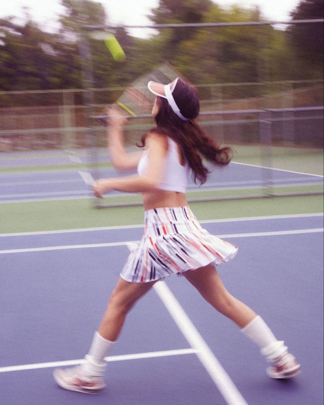 A female tennis player in a visor, white top, and skirt is mid-swing on an outdoor court, hitting a tennis ball with her racket.