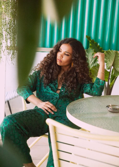 Woman in green patterned shirt and pants sitting at table with plants and green wall in background, looking away in relaxed pose.