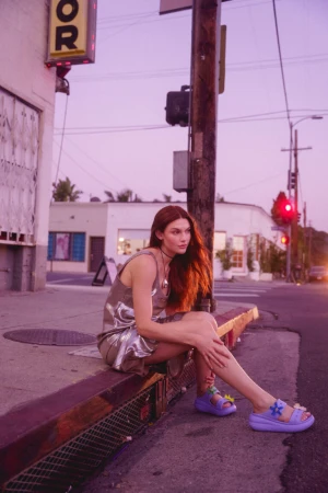 Woman in silver slip dress seated on a concrete curb next to a wooden utility pole under a purple sunset sky.