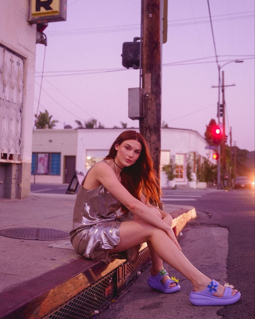 A woman with red hair sits on a curb on a city street, wearing a shiny dress and platform sandals at dusk, with city buildings in the background.