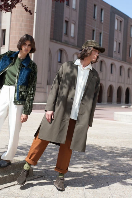 Two fashion models pose outdoors in stylish clothing. One wears a green jacket with white pants, the other a brown coat and trousers.