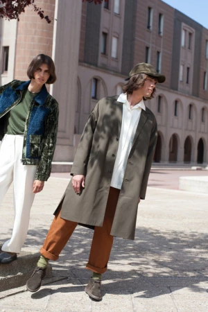 Two fashion models pose outdoors in stylish clothing. One wears a green jacket with white pants, the other a brown coat and trousers.