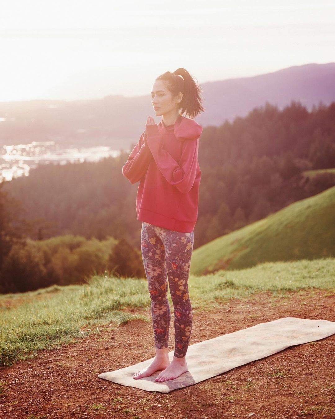 A slender woman practices yoga in an outdoor setting, wearing a reddish hoodie and floral pants, with mountains visible in the background.
