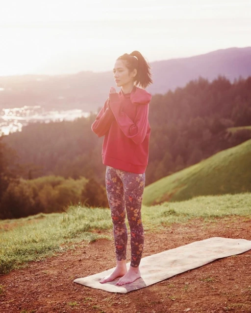 A slender woman practices yoga in an outdoor setting, wearing a reddish hoodie and floral pants, with mountains visible in the background.