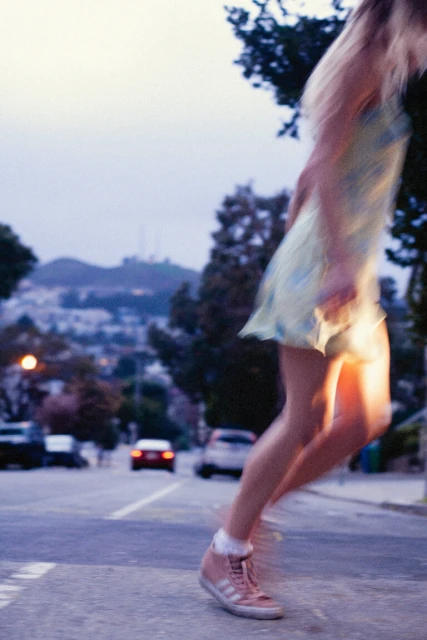 A blurred image of a woman in a light dress crossing a street in a residential neighborhood at dusk, with cars and houses visible.