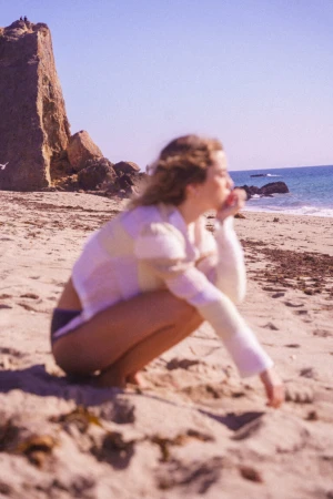 Woman in a light-coloured knitted top crouching on a sandy beach next to rocky outcrops and the ocean under a clear sky.