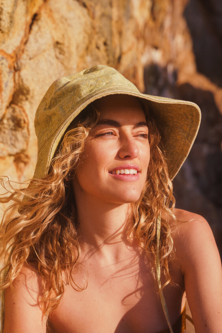 Portrait of an adult woman with curly hair, wearing a light-green hat, smiling in outdoor light with a rock formation background.