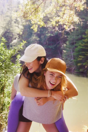 Two smiling young women wearing baseball caps outdoors in a wooded forest setting with a river behind them