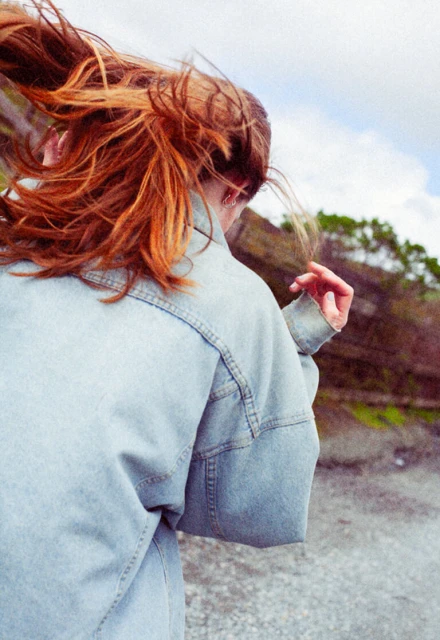 Woman with red hair wearing a denim jacket stands outdoors on a cloudy day. Trees and sky are visible in the background.