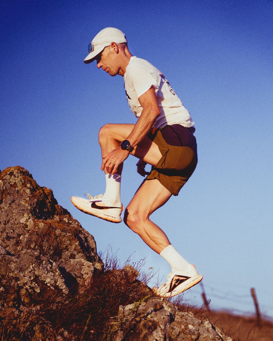 Athletic adult man in sportswear scaling rocks outdoors against a blue sky, showcasing fitness and nature photography.