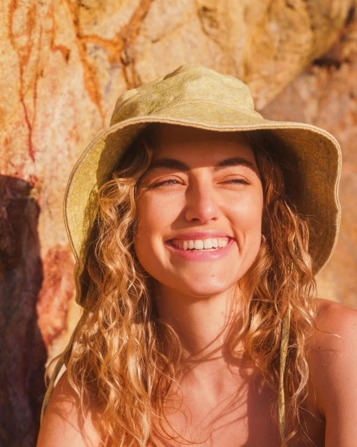 Portrait of a smiling woman with wavy blonde hair wearing a bucket hat with a rock formation in the background on a sunny day.