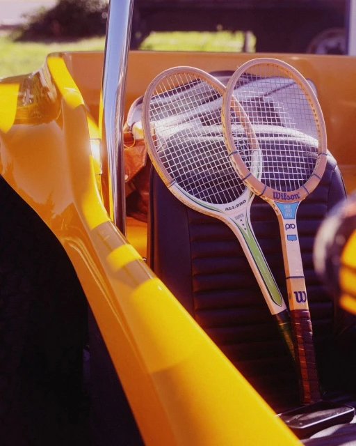 Two Wilson tennis rackets rest inside a yellow vehicle, bathed in sunlight. Close-up captures strings and logos against the dark interior.