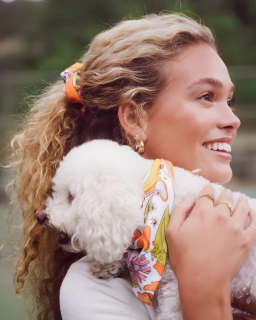 Blonde woman with curly hair smiles while holding a fluffy white dog wearing a patterned scarf in an outdoor setting on a sunny day.