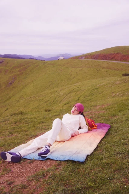 Woman relaxing outdoors on a colorful blanket in a grassy field. She wears a matching sweatsuit and hat with sneakers, enjoying the scenic view.