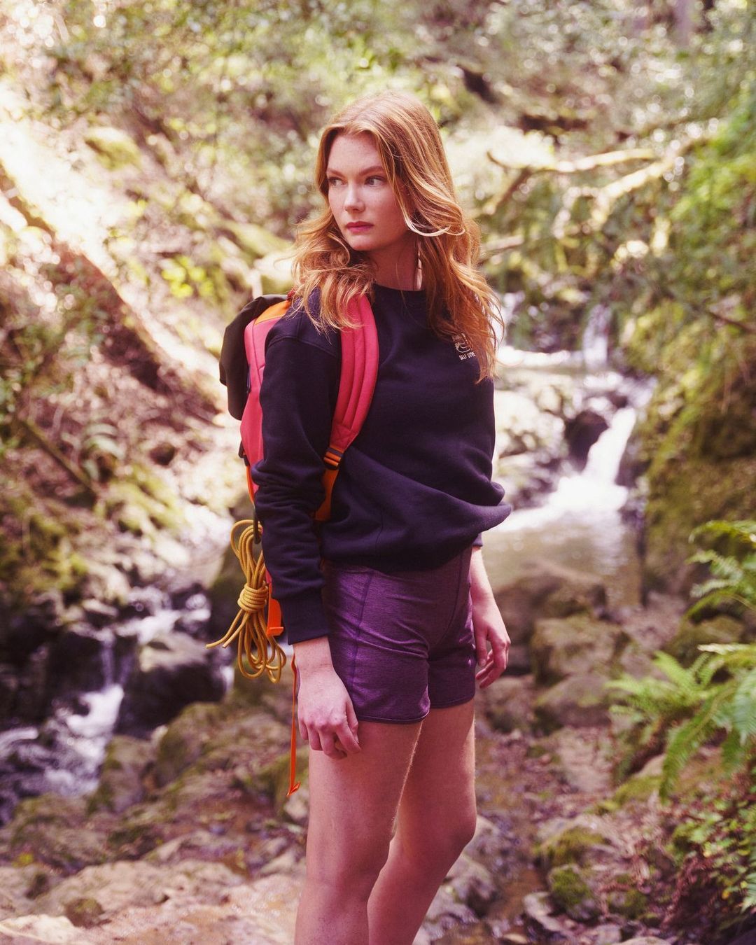 Red-haired woman wearing a dark sweatshirt and shorts with a backpack standing on a woodland trail.