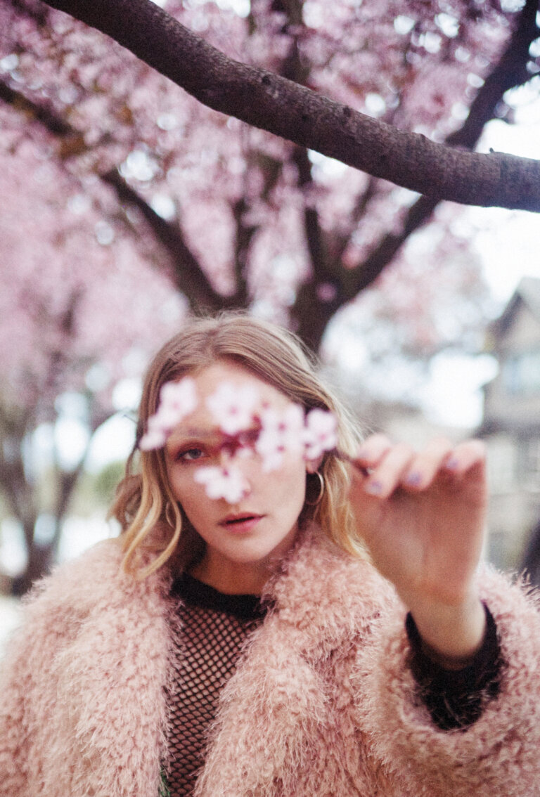 A woman stands outdoors in front of blossoming trees, holding a flowering branch in front of her face, wearing a fur coat.