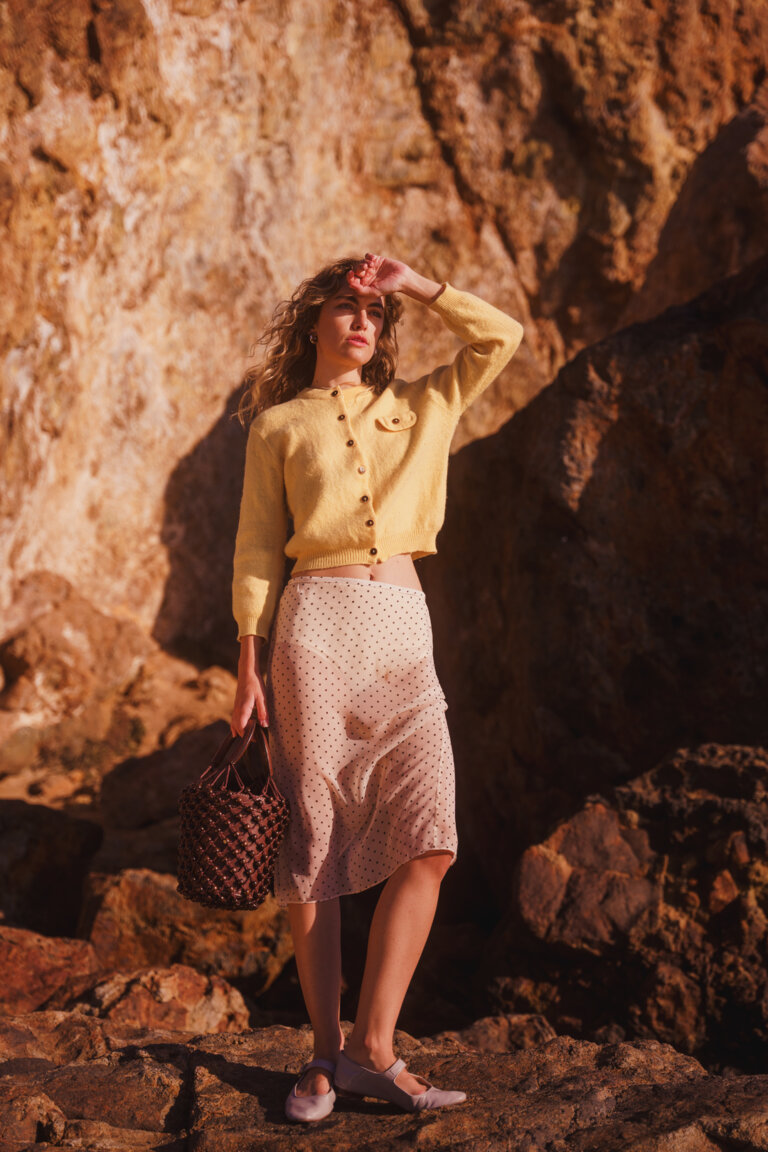 A woman in a yellow cardigan and polka dot skirt stands outside with a woven bag, posing against a rocky, sunlit backdrop.