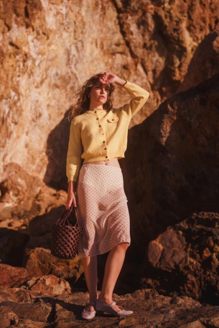 A woman in a yellow cardigan and polka dot skirt stands outside with a woven bag, posing against a rocky, sunlit backdrop.