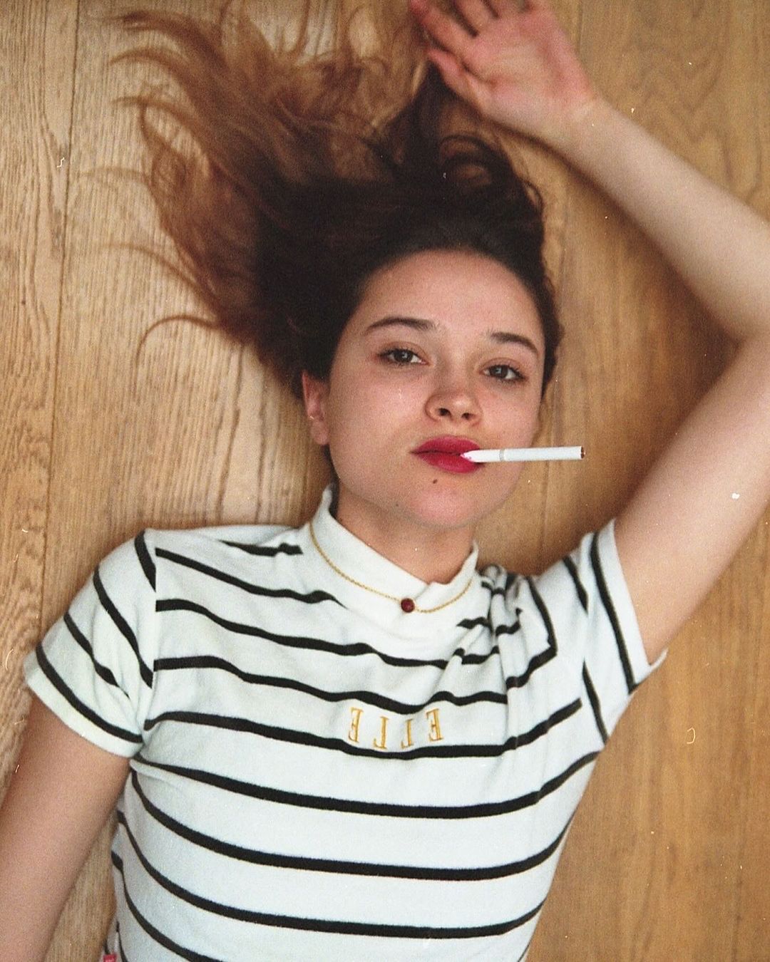 Woman with cigarette in mouth lies on wood floor. She wears a striped shirt with 'ELLE' text. Top-down view with indoor lighting.
