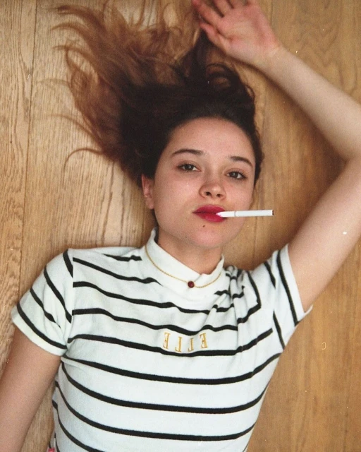 Woman with cigarette in mouth lies on wood floor. She wears a striped shirt with 'ELLE' text. Top-down view with indoor lighting.