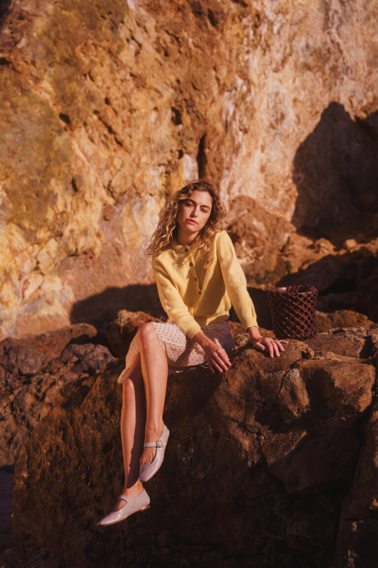 A woman with curly hair sits on a large rock in a yellow cardigan and patterned skirt. The background features a rocky cliff face.