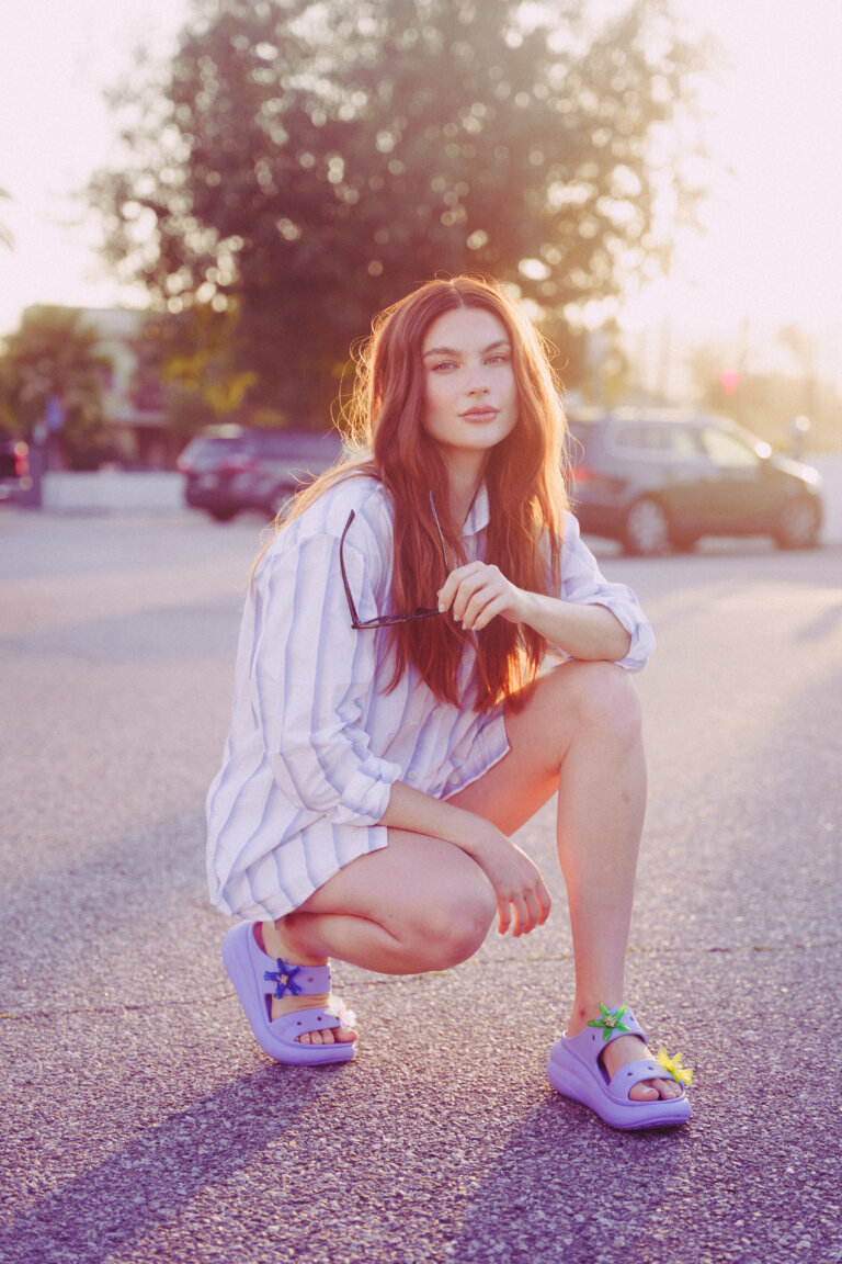 Red-haired woman in striped shirt and purple sandals crouching outdoors during golden hour by Nastasia Dusapin