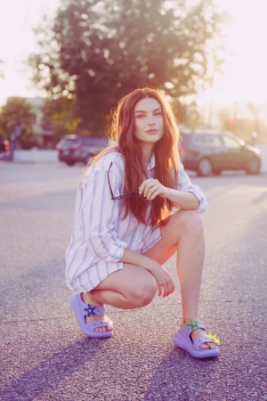 Red-haired woman in striped shirt and purple sandals crouching outdoors during golden hour by Nastasia Dusapin