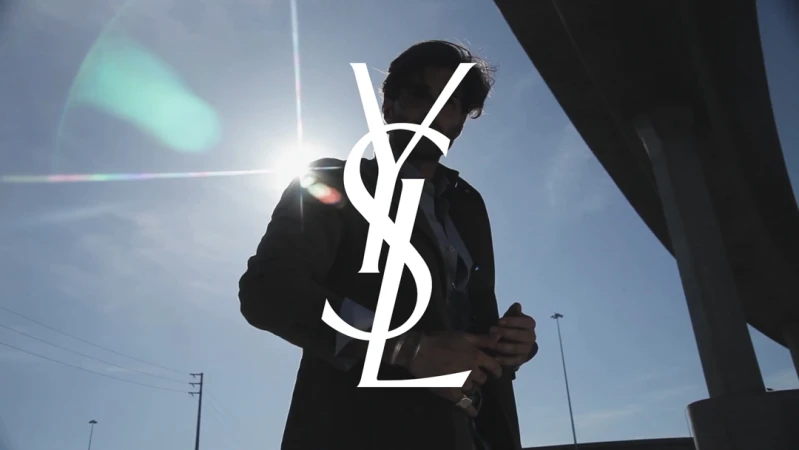 Low angle shot of a man standing outdoors against a bright sky with lens flare, featuring a large white YSL logo overlay.