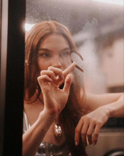 A young adult woman with red hair draws on a fogged glass pane with her finger, close up portrait, silver top, indoor setting.