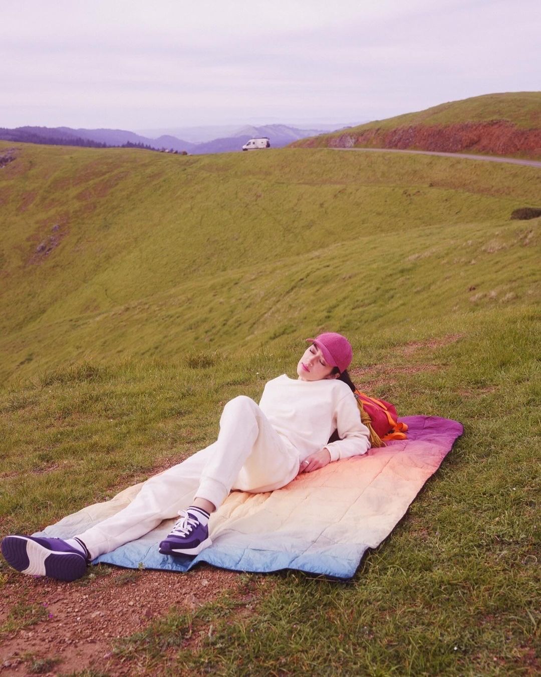 Woman in white sweatsuit relaxes on rainbow blanket in sunny grassy hills, wearing a red baseball cap, enjoying a scenic outdoor view.