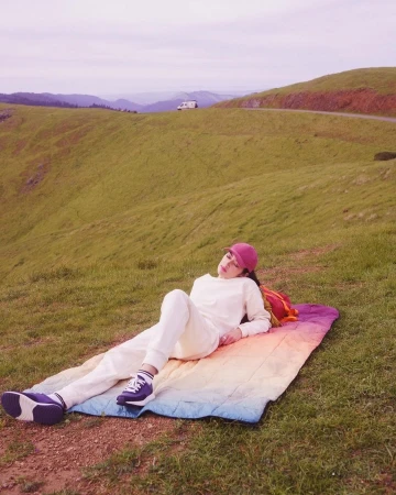 Woman in white sweatsuit relaxes on rainbow blanket in sunny grassy hills, wearing a red baseball cap, enjoying a scenic outdoor view.