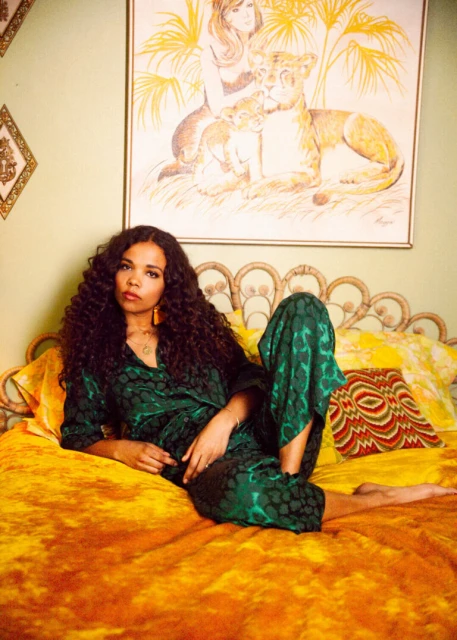 Adult woman with curly dark hair, wearing a green patterned pajama set, poses on a yellow and white patterned bed with a painting.