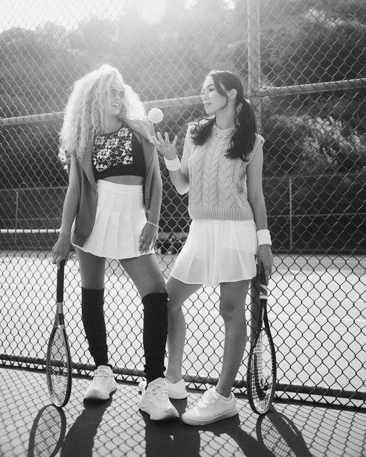 Two adult women in tennis outfits pose on a court next to a chain-link fence, one holding a ball, both with rackets.