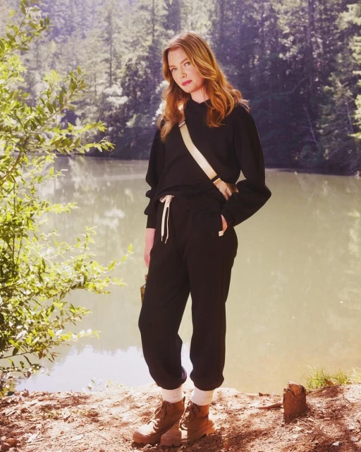 Woman in black sweatshirt and sweatpants poses outdoors near a lake surrounded by green trees, wearing hiking boots and carrying a bag.
