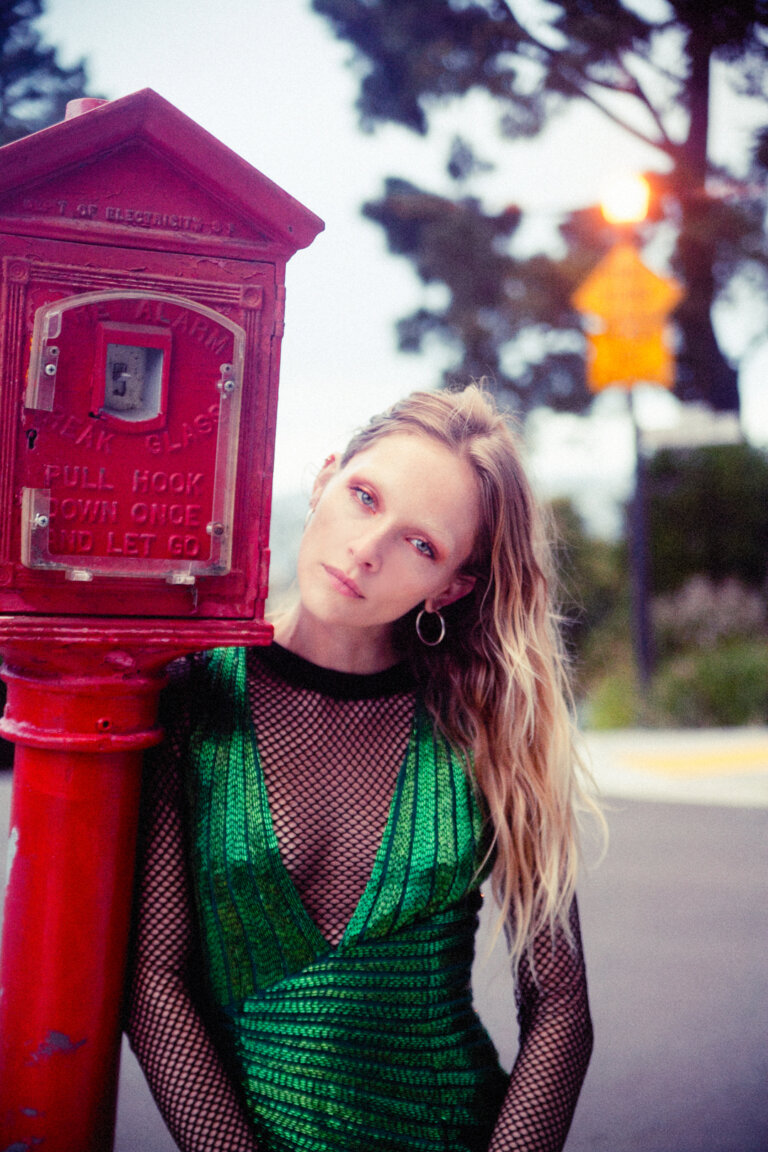 Woman in green dress poses by red alarm box. The scene is outdoors with trees in the background and the photo has a vintage feel.