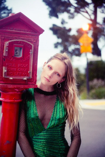 Woman in green dress poses by red alarm box. The scene is outdoors with trees in the background and the photo has a vintage feel.
