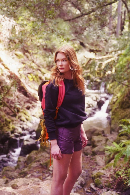 Adult female hiker with red hair wearing a backpack, shorts, and black sweatshirt poses in a nature setting with a small stream.