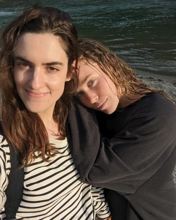 Two women posing together outdoors near water, one wearing a striped sweater and the other wearing a black sweatshirt