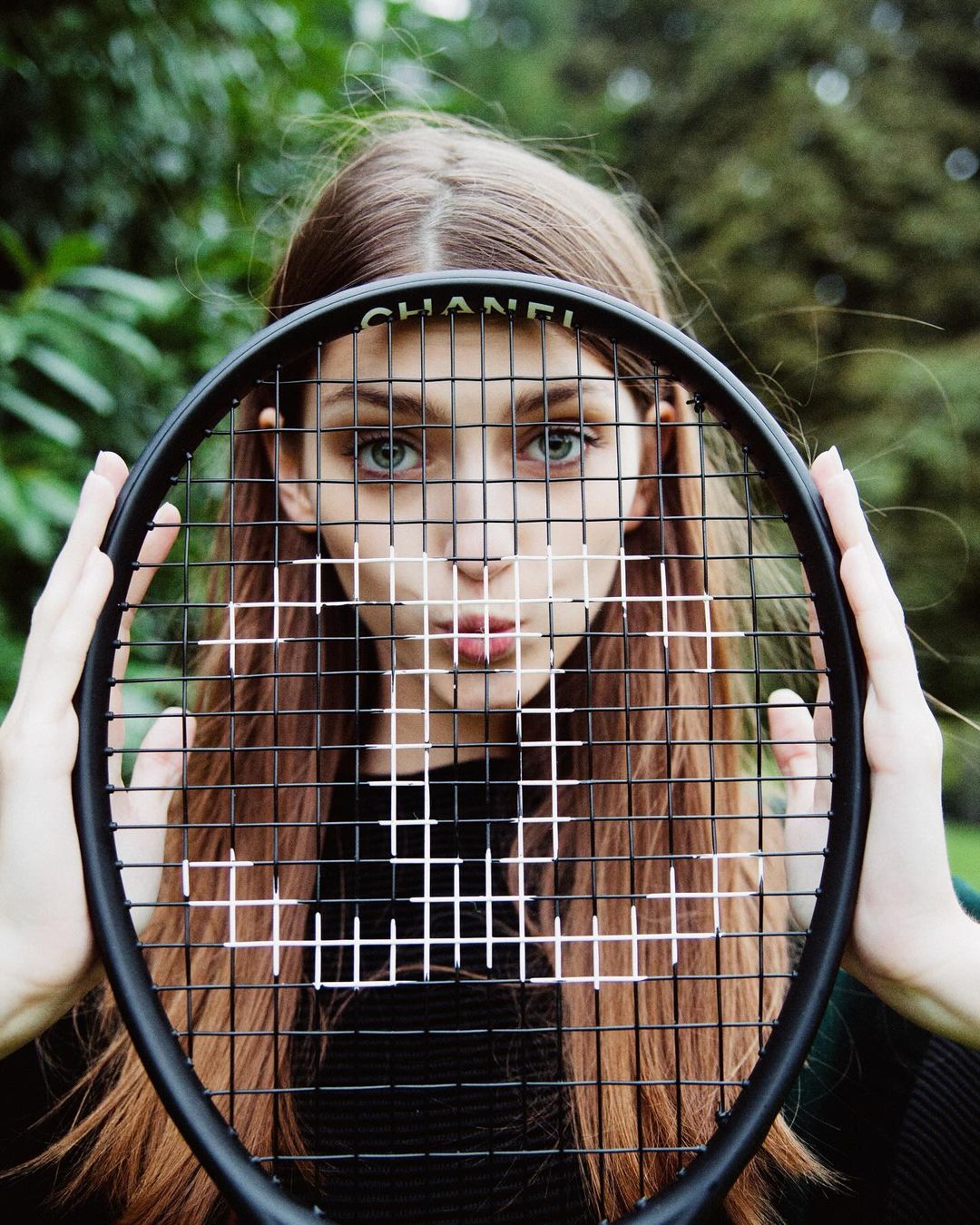 A woman is holding a Chanel-branded tennis racket over her face in an outdoor shot. Focus is on her eyes behind the racket's strings.