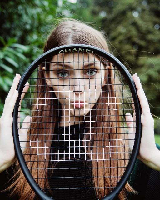 A woman is holding a Chanel-branded tennis racket over her face in an outdoor shot. Focus is on her eyes behind the racket's strings.