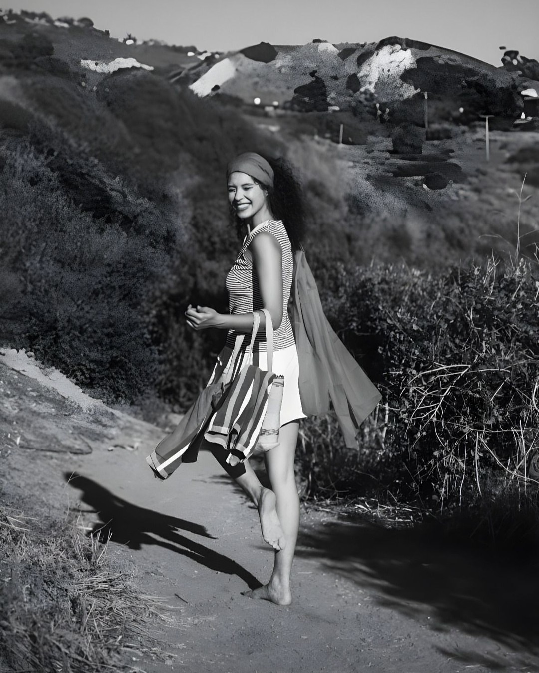 Woman walks on a path with a bag and shawl, smiling on a sunny day with green hills in the background. Black and white photograph.