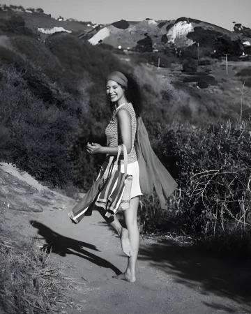 Woman walks on a path with a bag and shawl, smiling on a sunny day with green hills in the background. Black and white photograph.