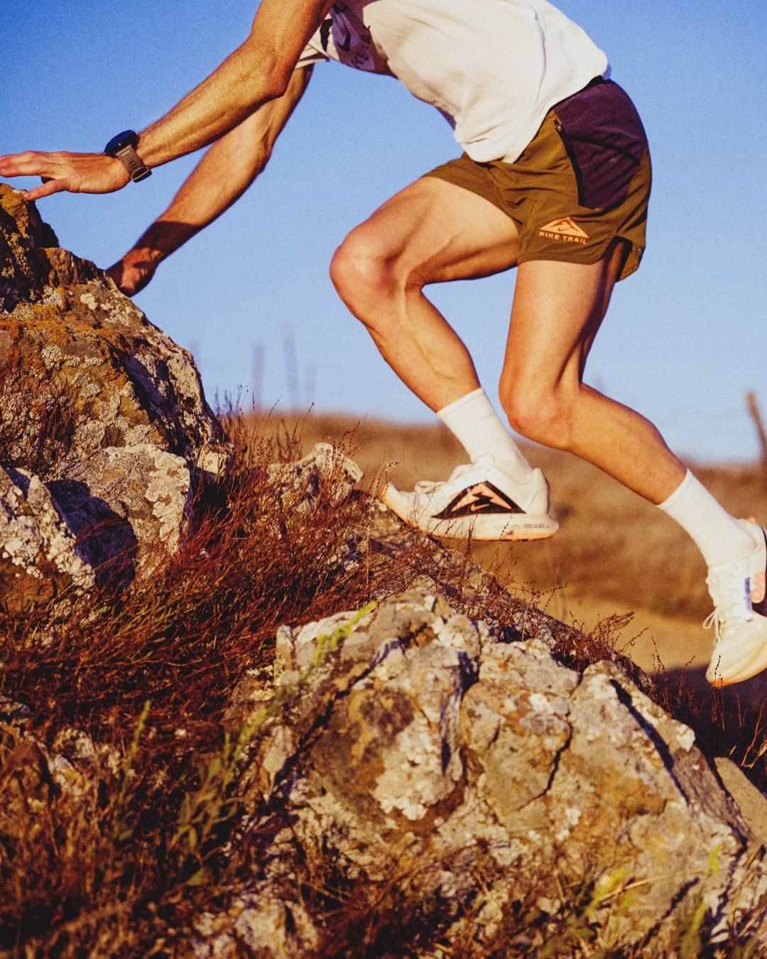 Trail runner climbs a rocky hill on a sunny day. The athlete wears running shoes, shorts, and socks while hiking through a dry landscape.