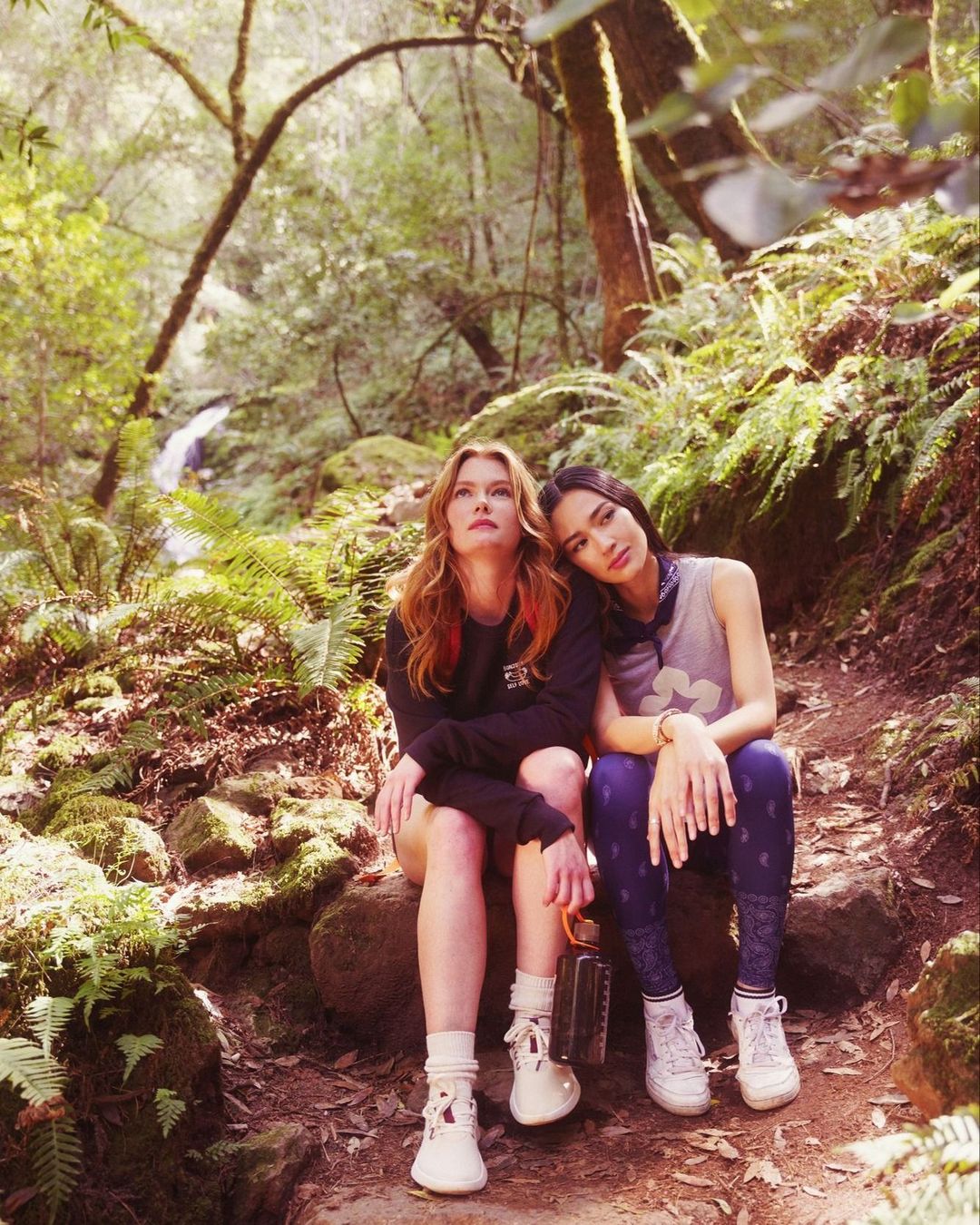 Two young women in outdoor fashion attire sit on a mossy trail in a sun-dappled forest, a waterfall visible in the background.