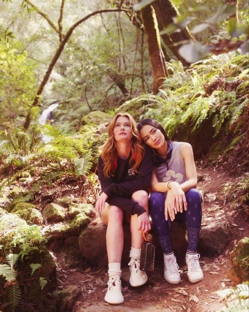 Two young women in outdoor fashion attire sit on a mossy trail in a sun-dappled forest, a waterfall visible in the background.