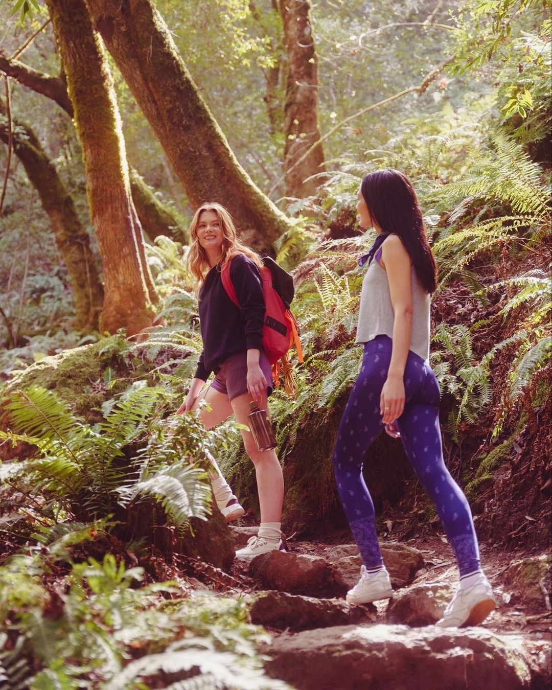 Two women are hiking on a sunny, fern-lined path in the woods, wearing athletic wear and carrying a water bottle and backpack.