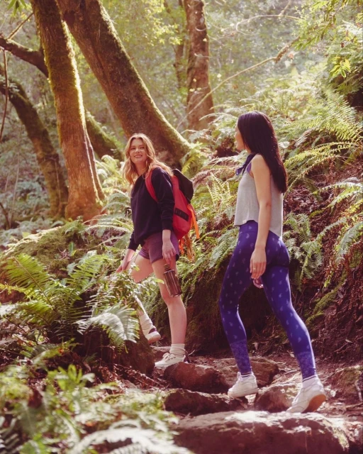 Two women are hiking on a sunny, fern-lined path in the woods, wearing athletic wear and carrying a water bottle and backpack.