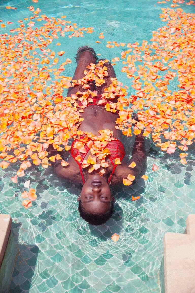 A dark-skinned woman wearing a red bikini floats on her back in a pool of clear water filled with orange rose petals, eye-level shot.