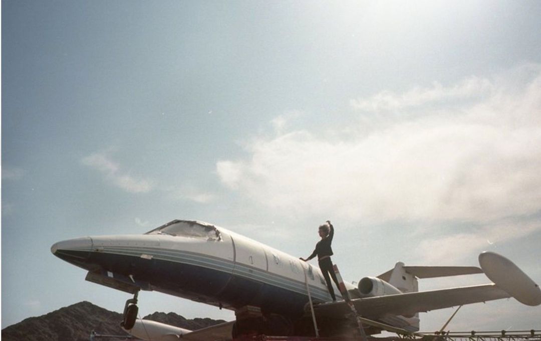 Adult stands atop private jet under a blue sky with white clouds. The person in a dark jumpsuit poses on the wing of the plane.